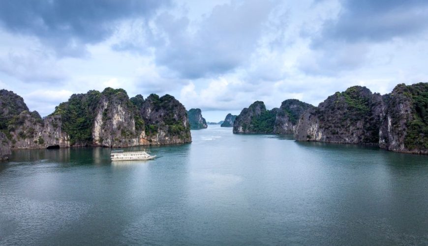 Aerial view of Halong Bay limestone karsts rising from emerald waters, with a cruise ship navigating between the islands