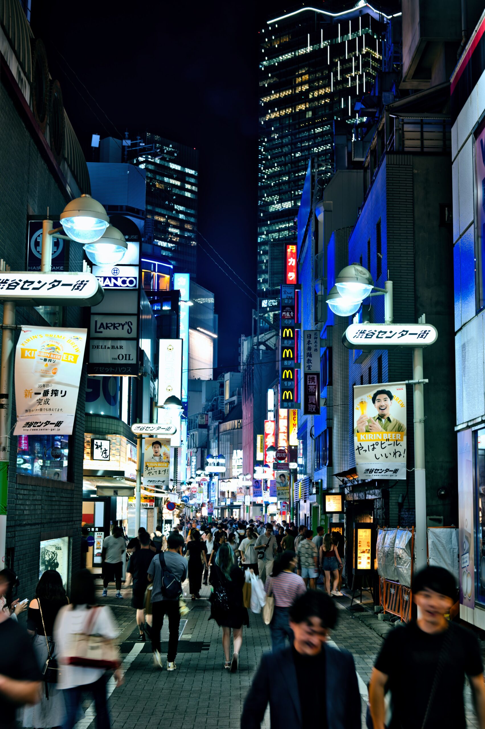 Pedestrians walk along Shibuya Center-gai at night past illuminated Japanese shop signs.