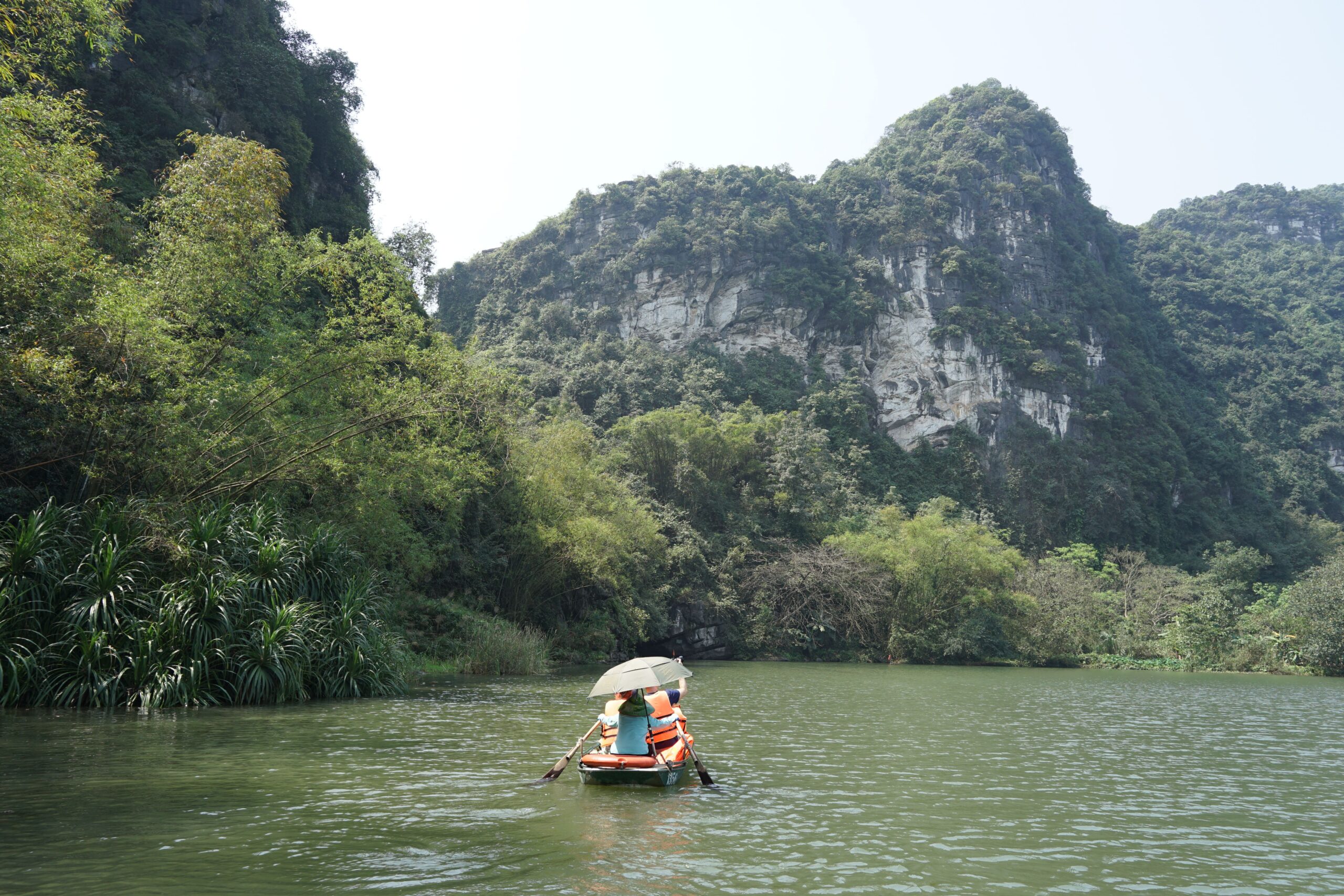 A covered visitor boat crosses a calm lake beneath steep green karst hills