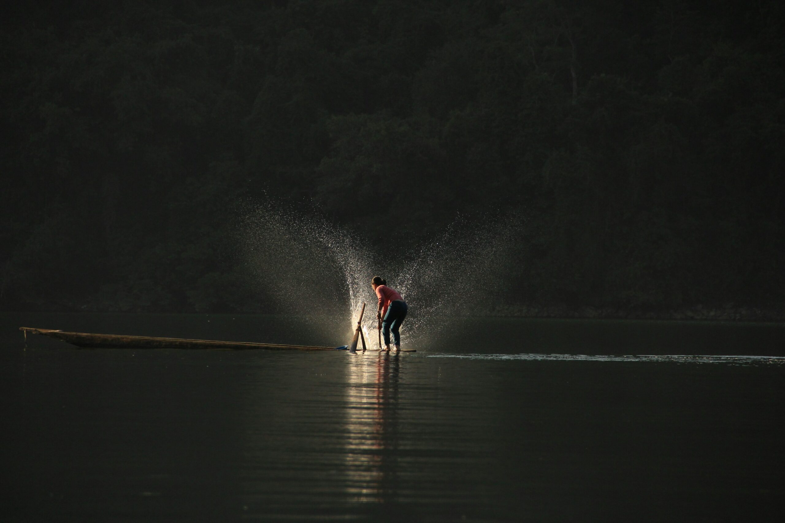 A fisher stands in shallow lake water beside a long wooden boat at dusk