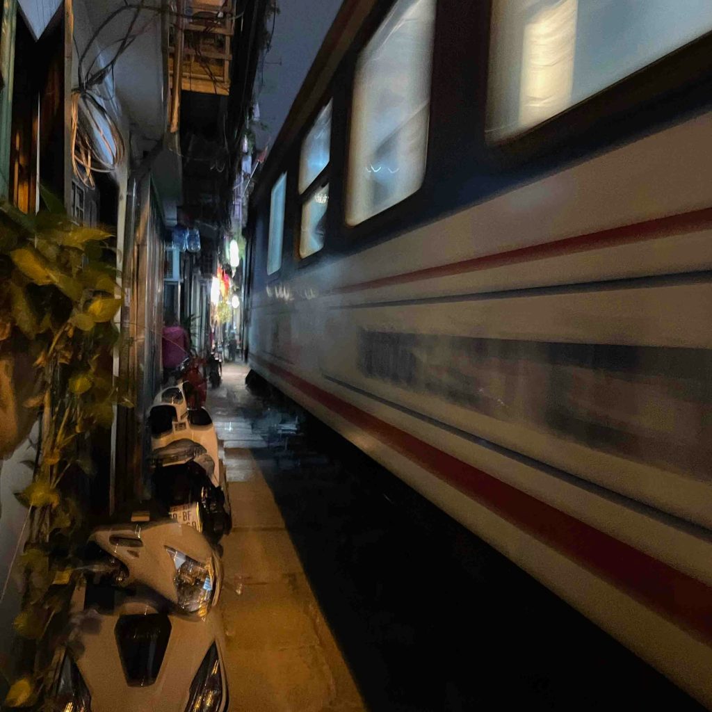 Passenger train passing through the narrow, illuminated Hanoi Train Street at night
