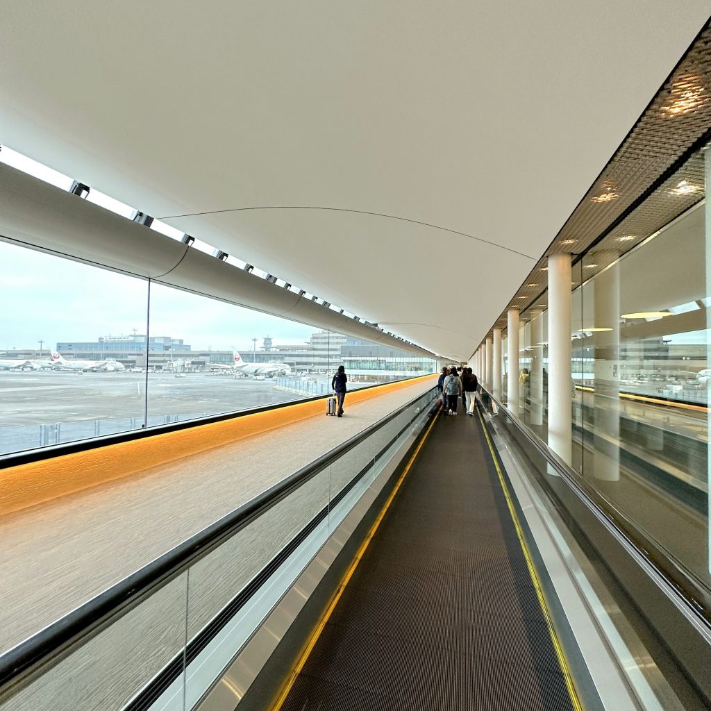 Travelers using a moving walkway inside a modern airport terminal overlooking parked aircraft