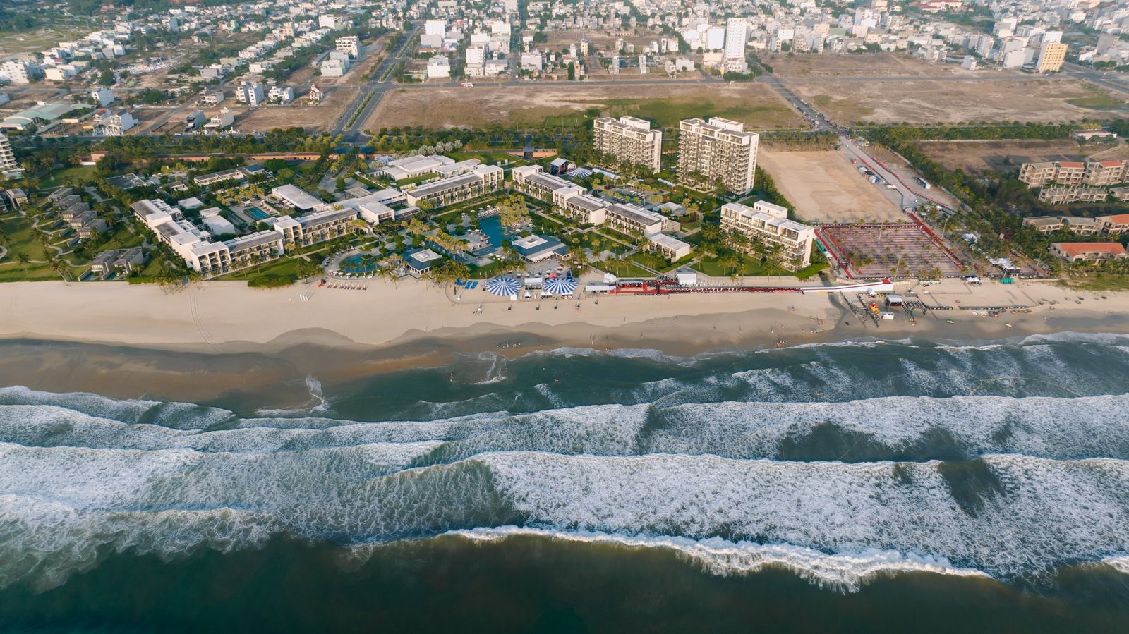 Aerial view of a beachfront resort with pools and villas set right along a sandy beach with ocean waves visible