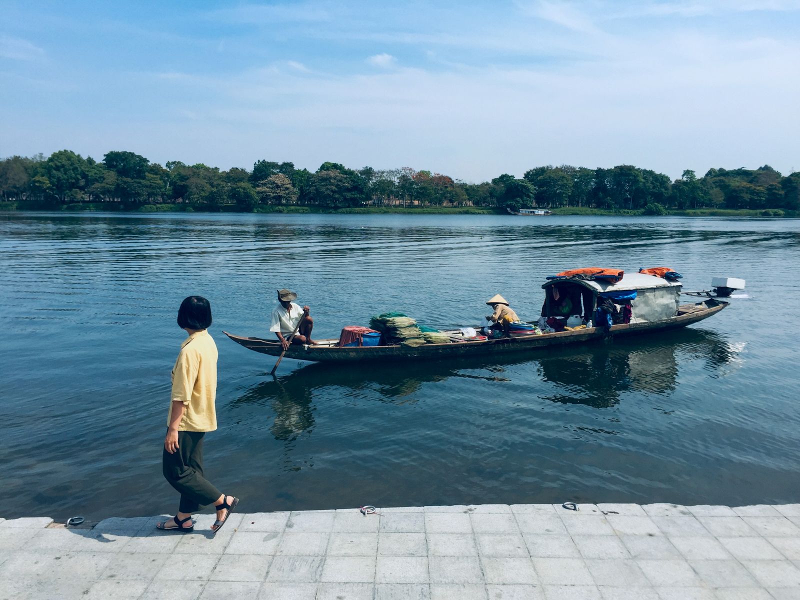 Traditional wooden boat with passengers and a nón lá hat gliding along the calm Perfume River in Hue, with a pedestrian walking on the tree-lined embankment