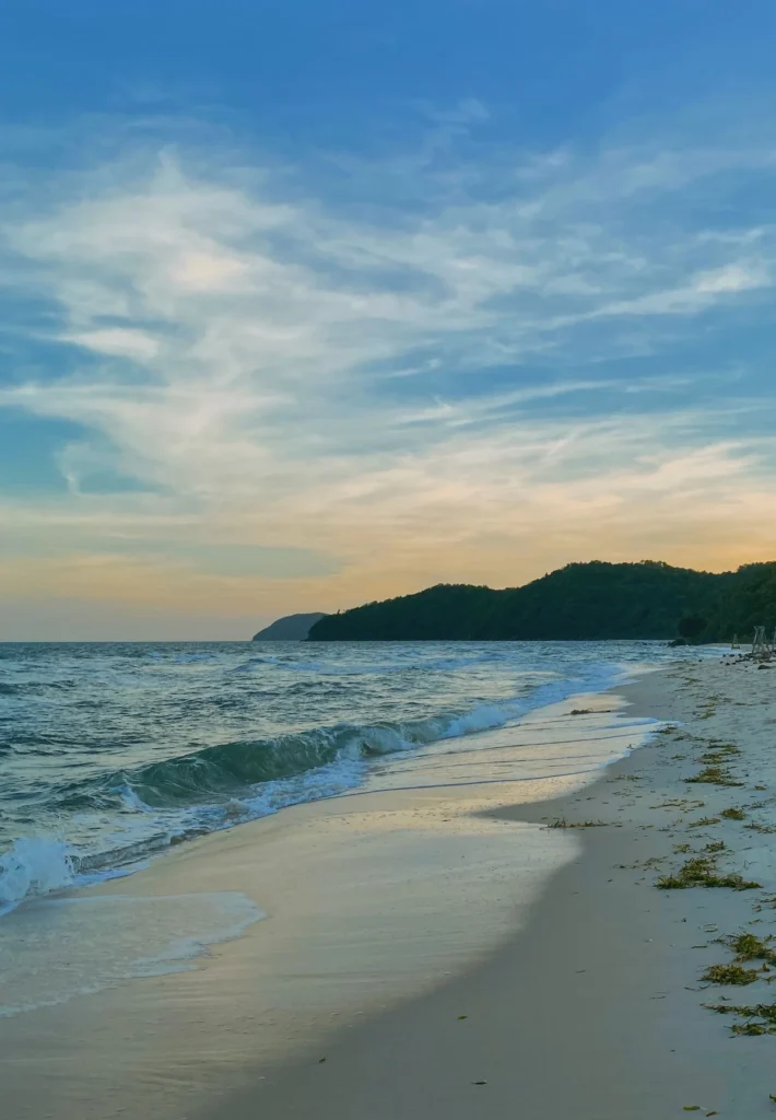 A Phu Quoc beach at sunset, with waves reaching the sand and a wooded headland beyond.