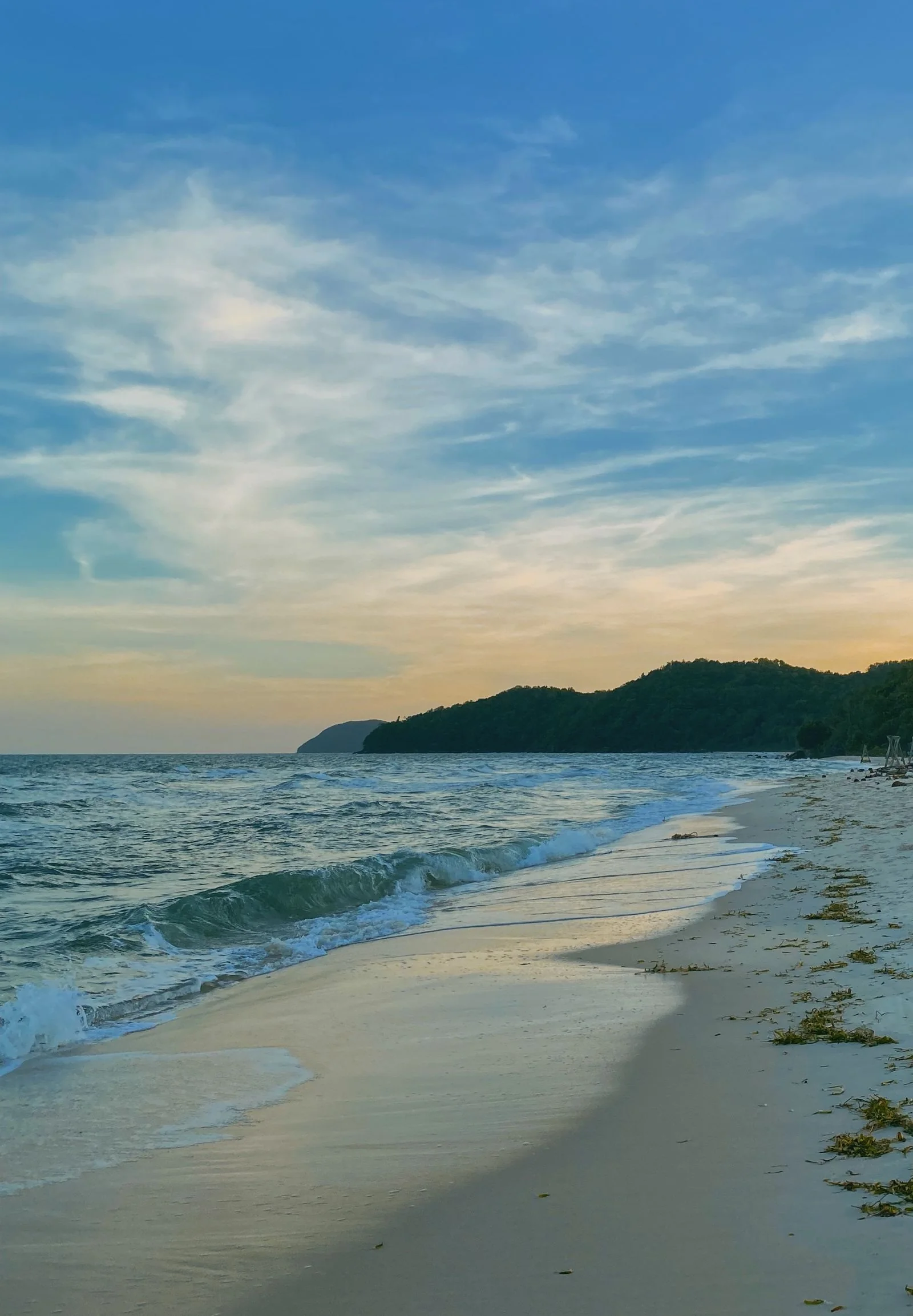 A Phu Quoc beach at sunset, with waves reaching the sand and a wooded headland beyond.